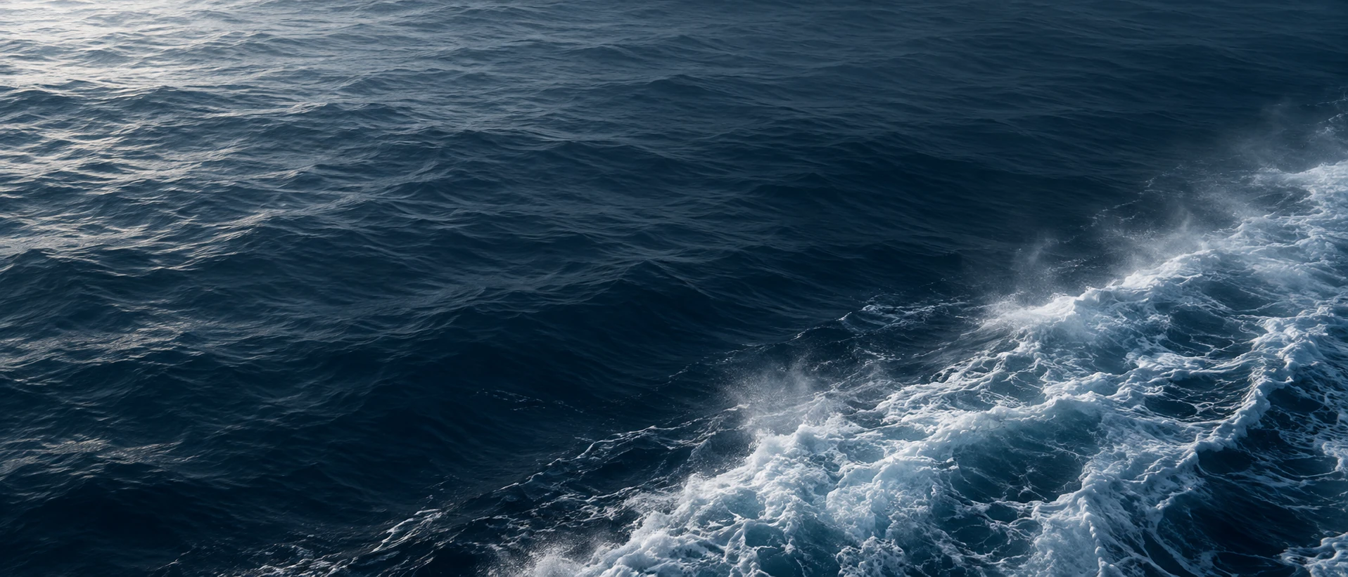 Minimal almost abstract macro ocean surface in ink blue with one soft diagonal trail of dissolving ship wake along the bottom right, upper and left areas calm, no ship visible.