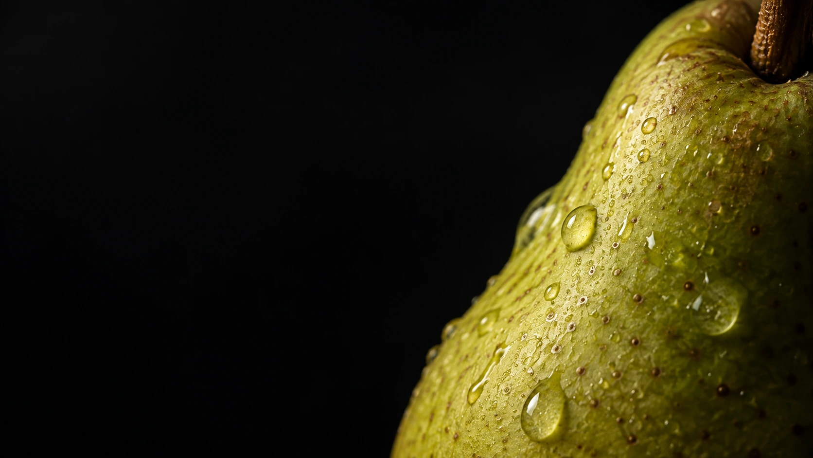 Extreme macro of the curved edge of a green pear at the right, tiny pores and a few natural water droplets visible, velvety black negative space.