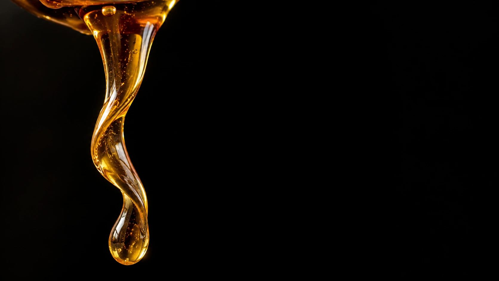 A slow ribbon of translucent amber honey suspended from an unseen source above, twisting narrowly on the right over a pitch-black backdrop.