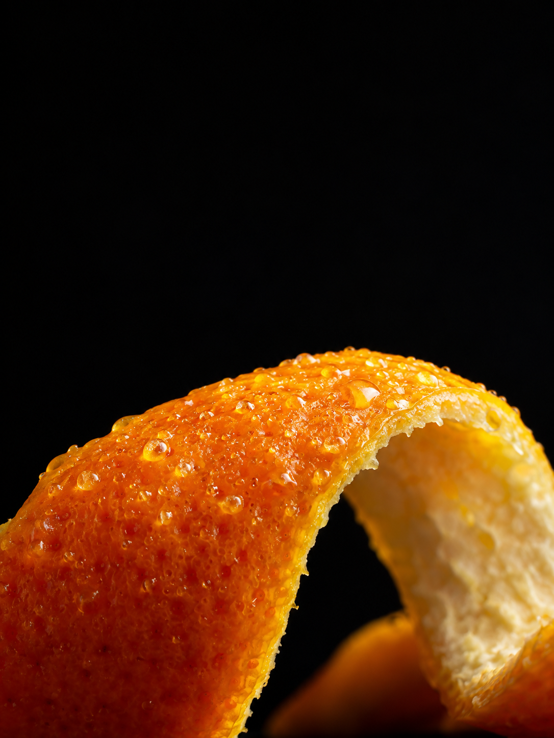 Ultra-close photographic view of a sharply curved fresh orange peel occupying the lower edge, oil glands glistening, black background.