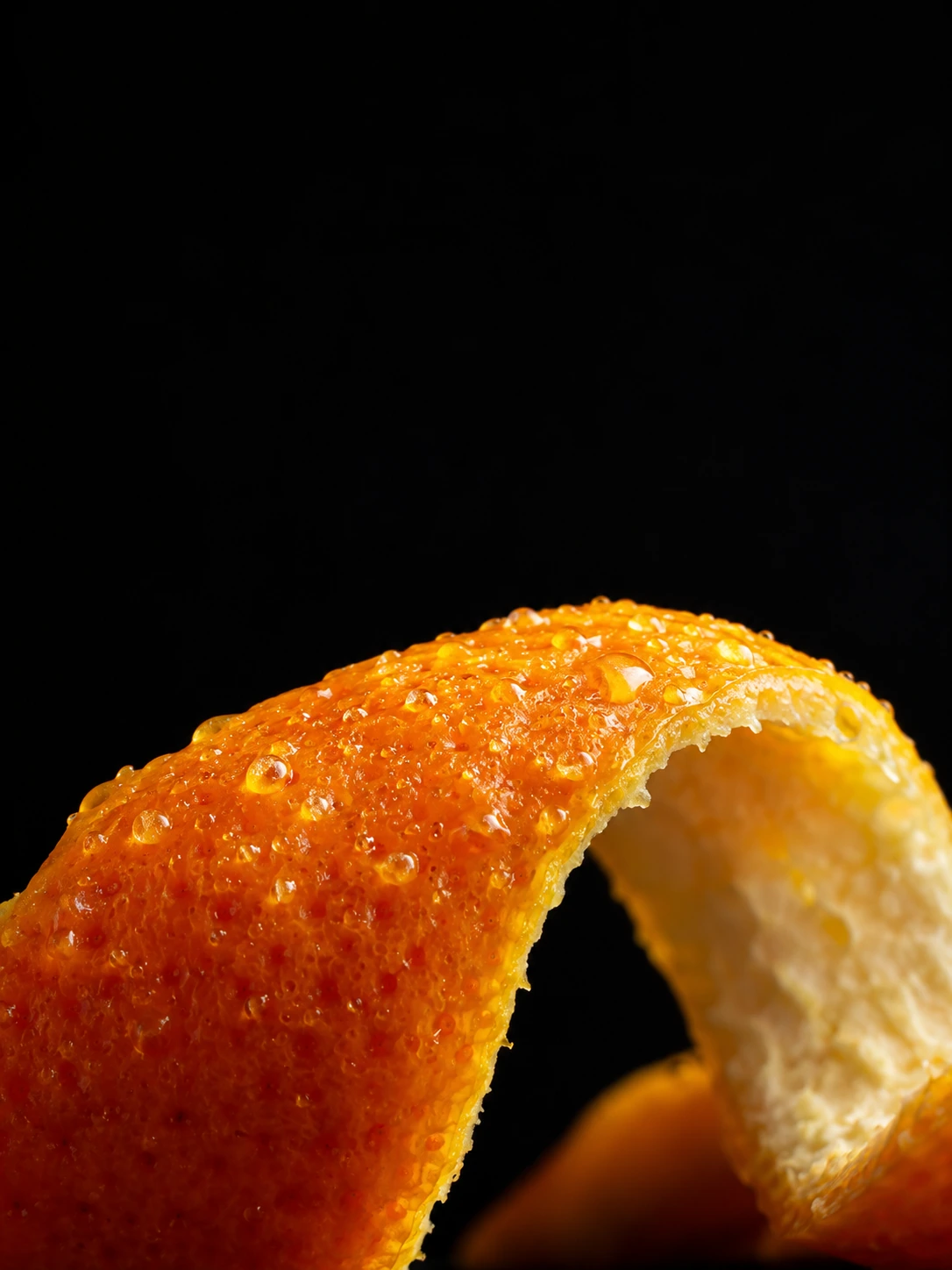 Ultra-close photographic view of a sharply curved fresh orange peel occupying the lower edge, oil glands glistening, black background.