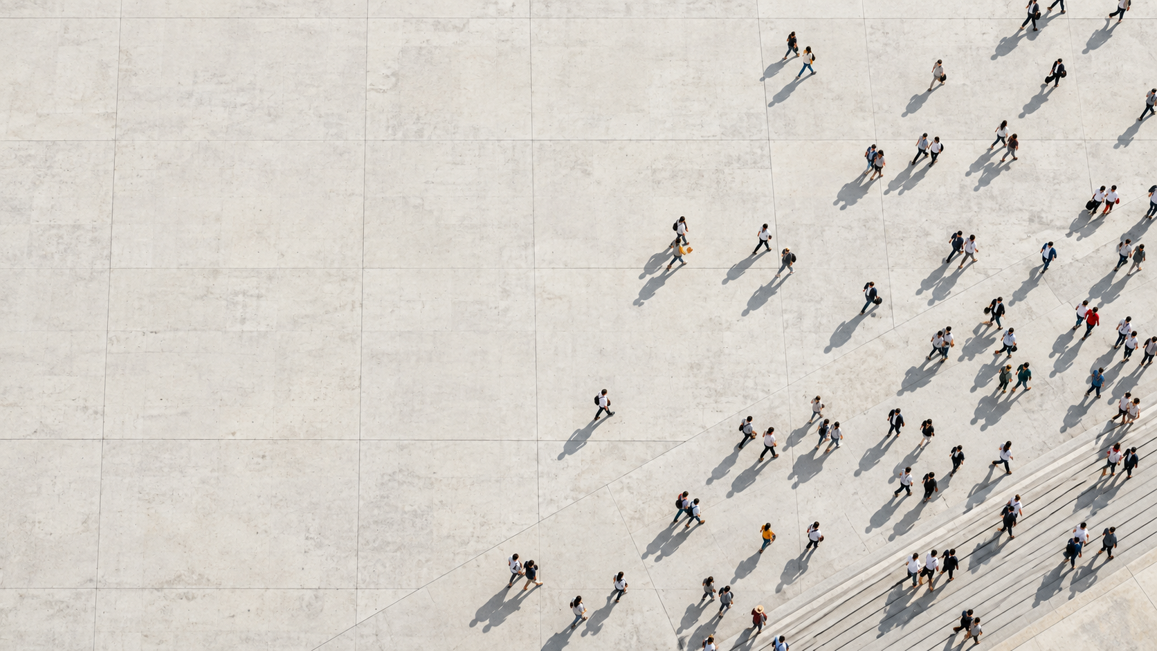 Direct overhead view of many tiny adult pedestrians crossing a huge pale concrete plaza in loose natural paths, activity concentrated along the lower right.