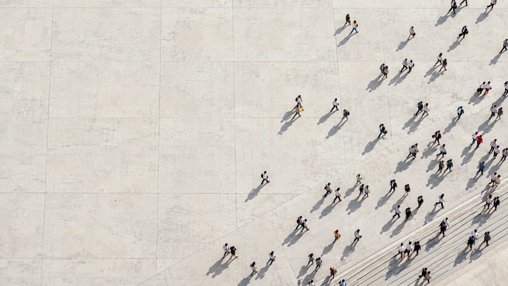 Direct overhead view of many tiny adult pedestrians crossing a huge pale concrete plaza in loose natural paths, activity concentrated along the lower right.