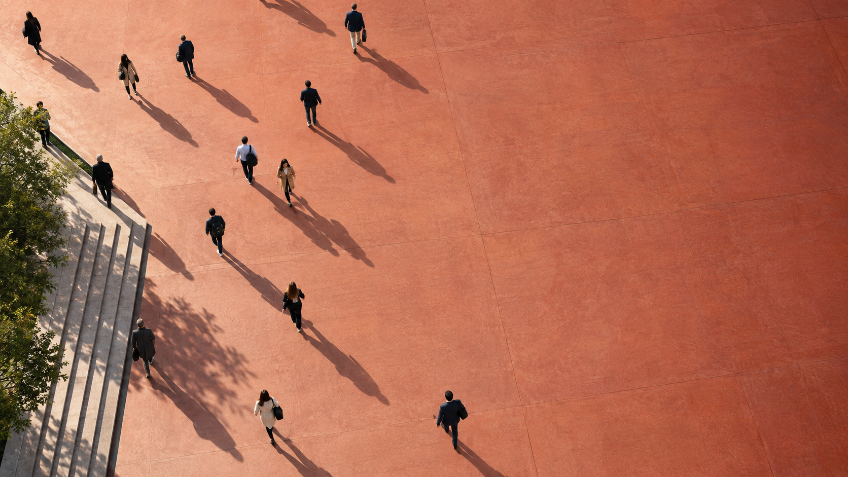 Aerial view of pedestrians walking across a broad terracotta-red urban promenade, long morning shadows, a large open area on the left.