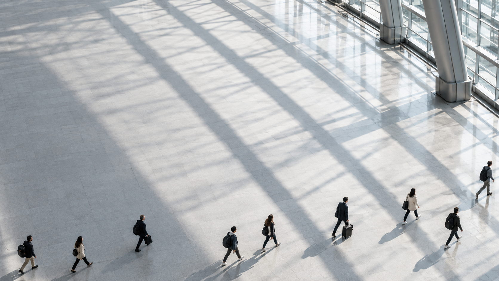 High-angle view of small commuters spaced along the lower edge of a vast clean station concourse, silver roof shadows forming broad diagonal lines.