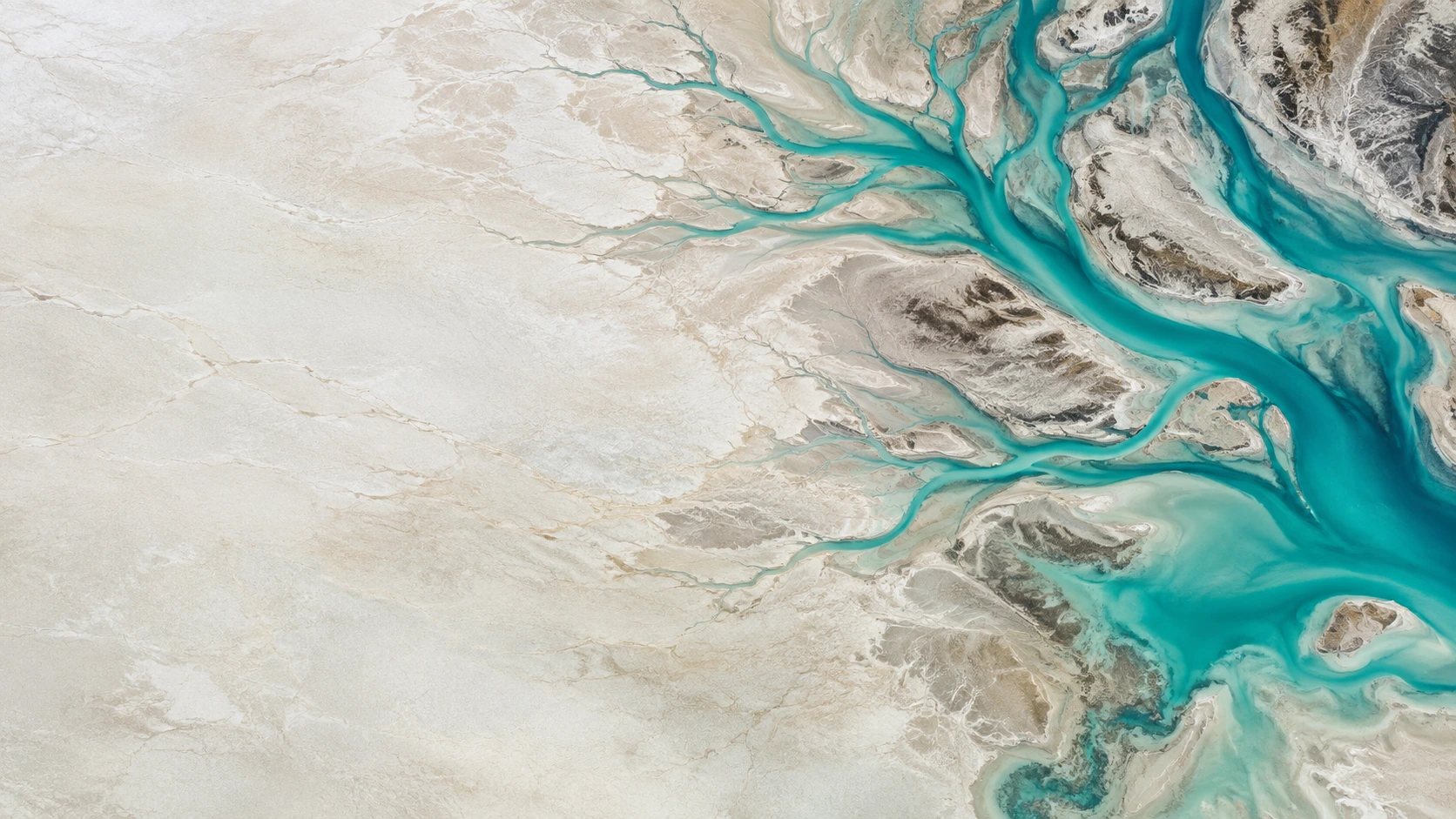 Overhead aerial of delicate turquoise tidal channels branching through a vast ivory salt flat, channels clustered toward the right.