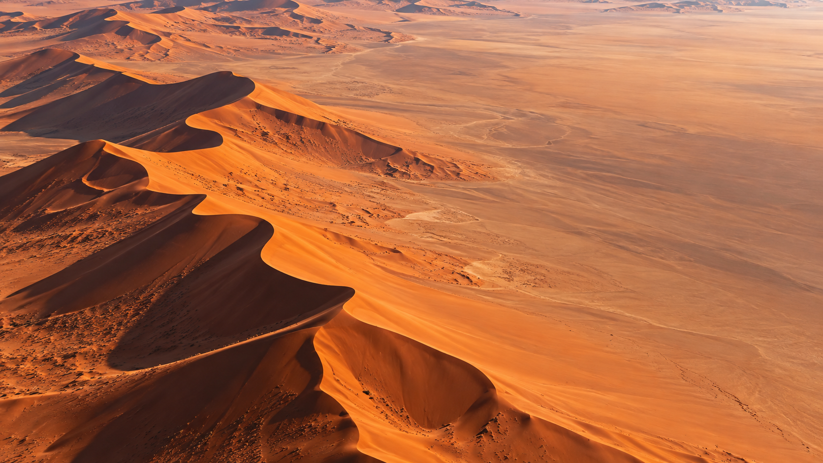 High aerial view of large red-orange desert dunes with razor-sharp ridges and deep long shadows, clean expansive upper sand.