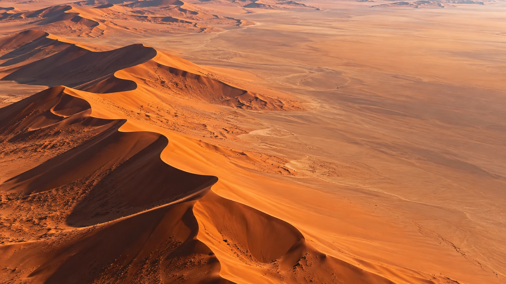 High aerial view of large red-orange desert dunes with razor-sharp ridges and deep long shadows, clean expansive upper sand.