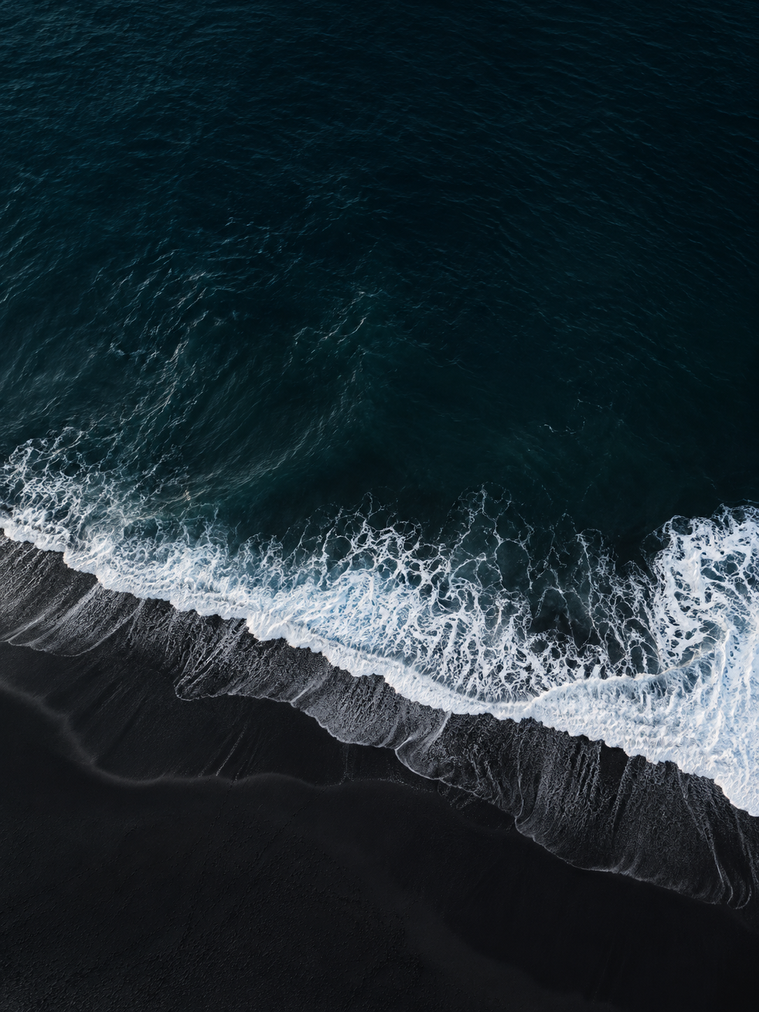 Aerial overhead of a single fine white wave breaking across pitch-black volcanic sand, minimal almost graphic composition.