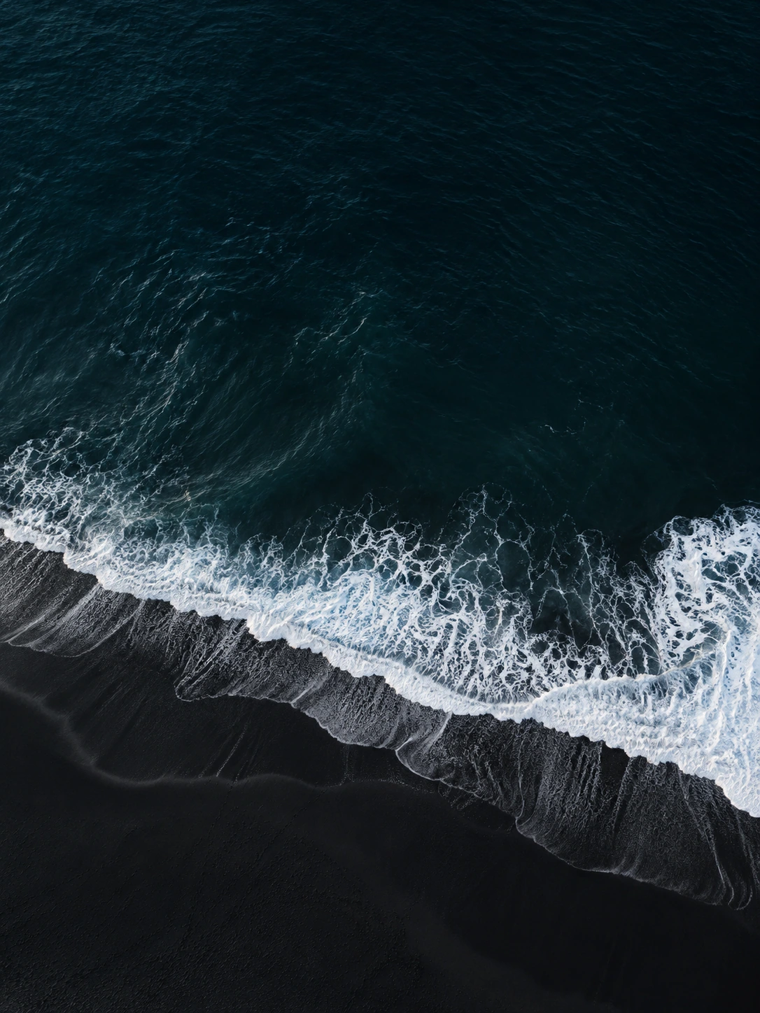 Aerial overhead of a single fine white wave breaking across pitch-black volcanic sand, minimal almost graphic composition.