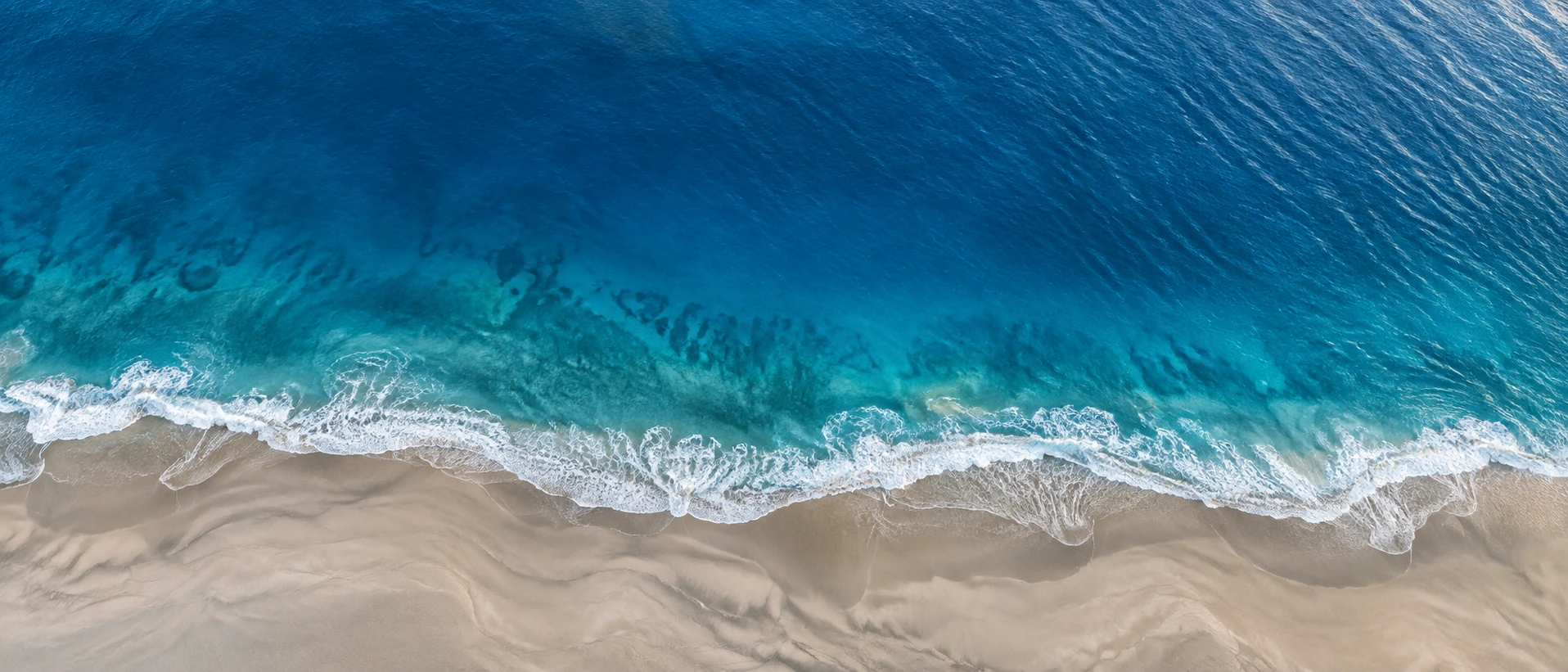 Panoramic high-altitude view of a thin foamy white coastline dividing muted sand below from expansive clear blue sea above.