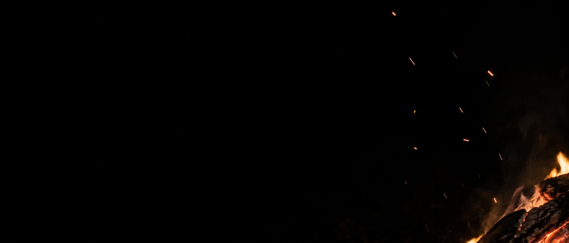 A few uneven amber sparks rise above a cropped campfire at the lower right.