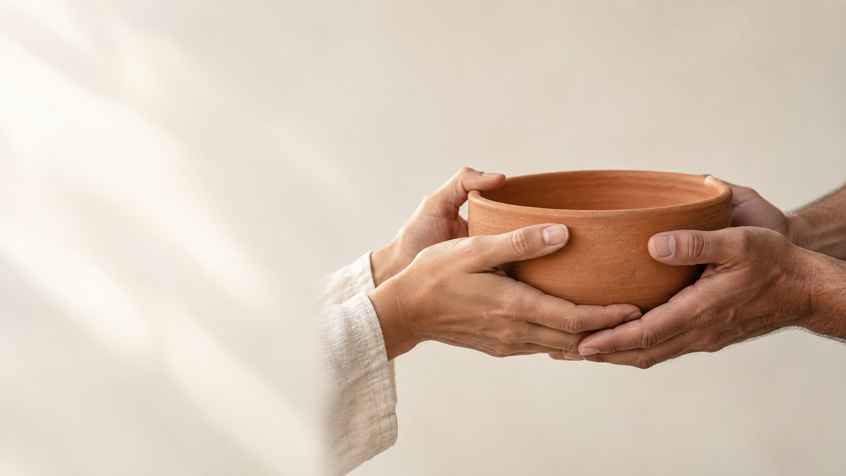 Close-up of two adults' hands jointly holding one plain terracotta bowl at its opposite sides, natural skin texture, ivory studio background.