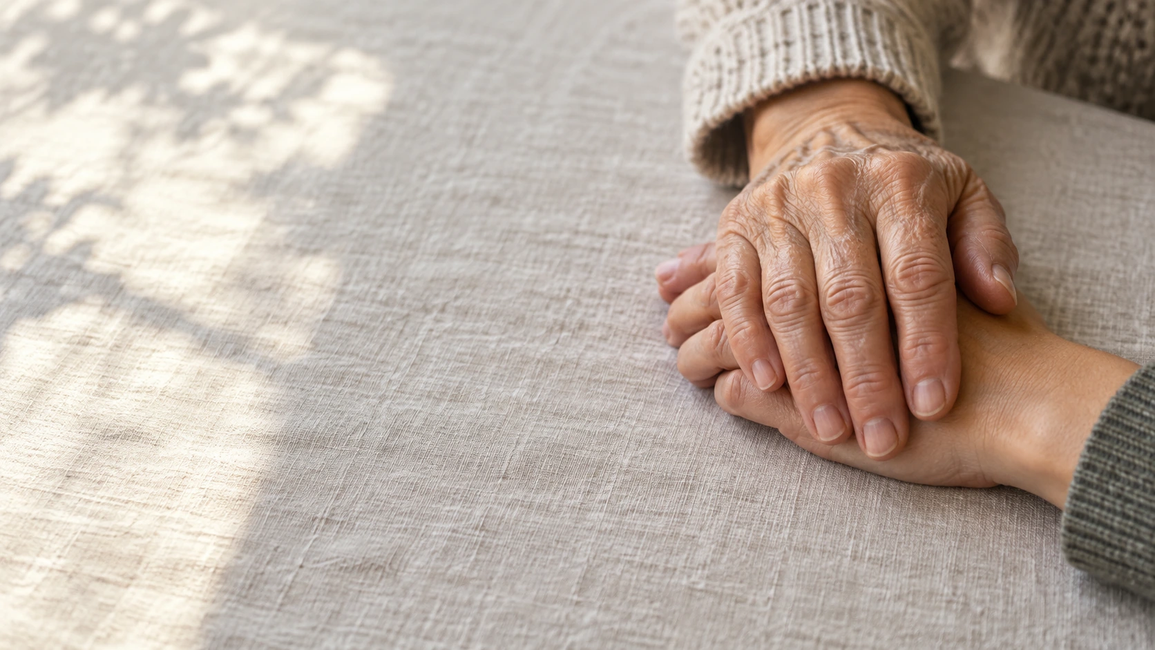 Close-up of one older adult hand resting gently over one younger adult hand on a neutral linen table, intimate documentary realism.